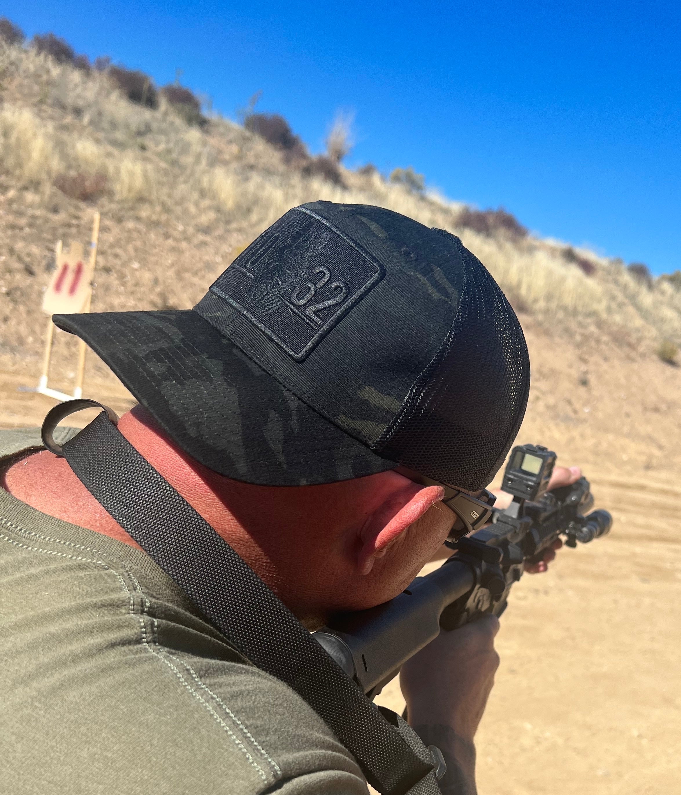 a person holding a rifle at a shooting range with a clear blue sky and desert landscape wearing a 10-32 Stealth lid.